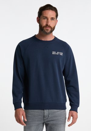 Sötétkék crewneck pulóver raglán ujjal, texturált anyaggal és fehér felirattal a mellrészen. Világosszürke farmernadrággal párosítva.