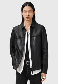 Veste en cuir noir avec un design à col, fermeture éclair à l'avant et deux poches zippées. Texture lisse et style minimaliste complètent une tenue superposée.