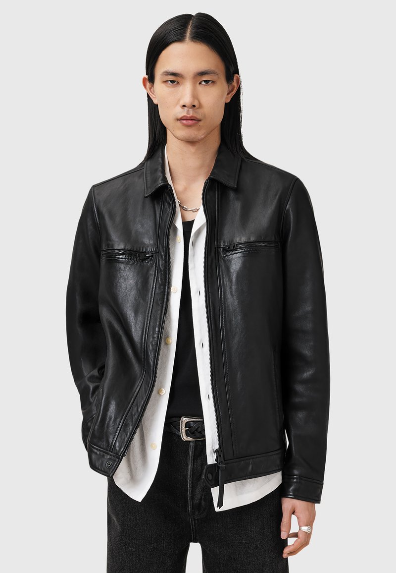 Veste en cuir noir avec un design à col, fermeture éclair à l'avant et deux poches zippées. Texture lisse et style minimaliste complètent une tenue superposée.