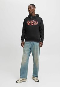 Schwarzer Hoodie mit "Stranger Things" Grafik in orange-rot flammendem Design, kombiniert mit verwaschenen blauen Jeans und beigen Turnschuhen.