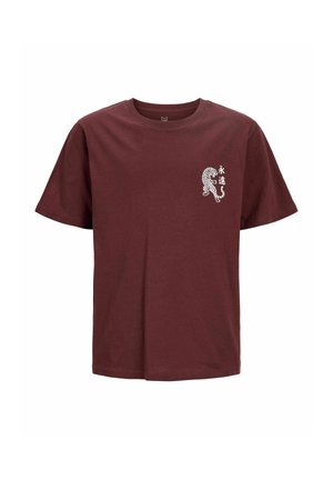 Młody mężczyzna uśmiecha się, ma na sobie bordowy t-shirt z małą grafiką tygrysa, luźne czarne dżinsy i beżowe sneakersy, stoi na jednolitym tle.