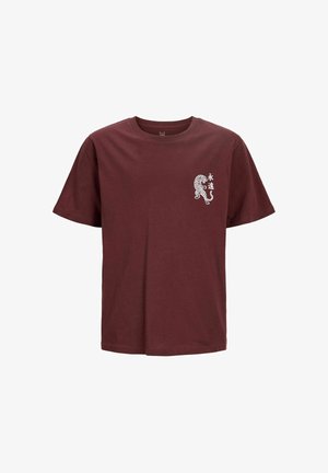 Młody mężczyzna uśmiecha się, ma na sobie bordowy t-shirt z małą grafiką tygrysa, luźne czarne dżinsy i beżowe sneakersy, stoi na jednolitym tle.