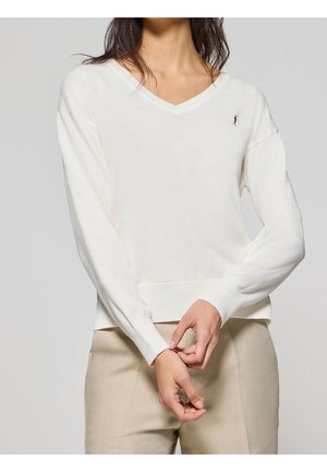 Femme portant un pull blanc à col en V à manches longues avec un petit logo brodé, associé à un pantalon beige, les mains jointes devant.
