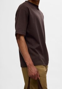 Camiseta de algodón marrón de manga corta, con cuello redondo y corte relajado, acompañada de pantalones beige; mano sosteniendo un bolso marrón oscuro.