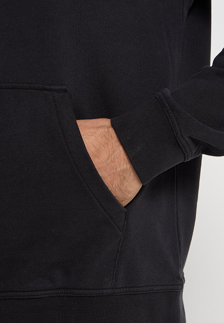 Czarny sweatshirt zaprezentowany z ręką w przedniej kieszeni. Posiada ściągacze przy mankietach oraz miękkie, teksturowane wykończenie tkaniny.