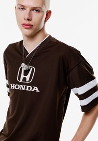 Bruine korte mouwen jersey met witte strepen, voorzien van een prominent Honda-logo. Soepele textuur met een v-hals ontwerp. Zilveren kettingdetail.