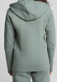 Capuchontrui in lichtgroen, gemaakt van gestructureerde stof. Heeft een getailleerd ontwerp met lange mouwen en een klein logo bij de zoom.