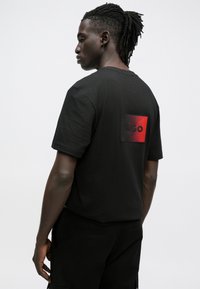 Μαύρο βαμβακερό t-shirt με ορθογώνιο κόκκινο και μαύρο λογότυπο με την ένδειξη "HUGO" στο πάνω μέρος της πλάτης, με κοντά μανίκια και άνετη εφαρμογή.