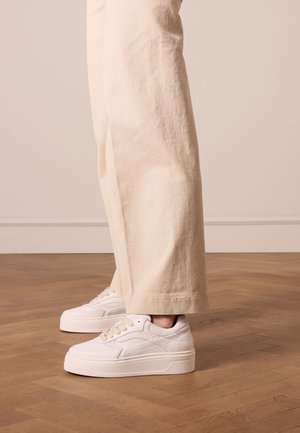 White platform sneakers with smooth leather upper, subtle textured accents, and beige laces, paired with wide-leg cream pants.