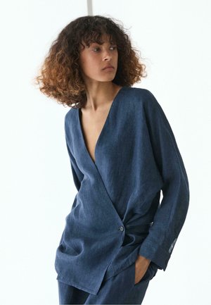 Frau mit lockigem Haar, die einen dunkelblauen Wickel-Blazer mit tiefem V-Ausschnitt und einem Knopf trägt, steht mit der Hand in der Tasche und schaut seitlich.