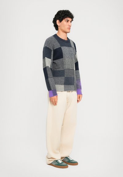 Hombre de pie de lado, vistiendo un suéter de parches azul grisáceo, pantalones holgados beiges y zapatillas verdes con suelas marrones.