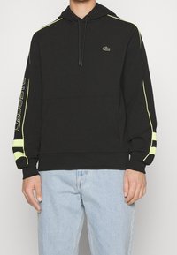 Svart hoodie med gula accentstripes på axlarna och ärmarna, med en framficka och dragsko i huvan. Lacoste-logotypen finns.