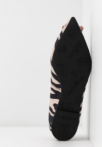 Chaussure plate à bout pointu avec dessus à imprimé zèbre, montrant une semelle noire avec marquage embossé et petites icônes de chaussures, appuyée contre des murs blancs.