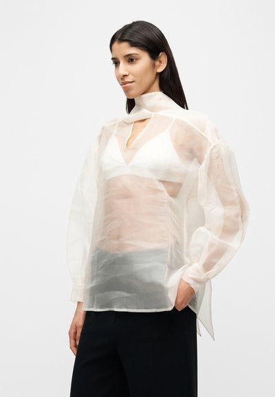 Λευκό διάφανο μπλουζάκι με ψηλό λαιμό και λεπτομέρεια με άνοιγμα. Φουσκωτά μανίκια και ελαφρώς δομημένη σιλουέτα που προσφέρουν υφή.