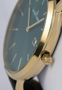 Goldfarbene Uhr mit grünem Ziffernblatt, schlankem Profil und einfachen Stundenmarkierungen. Verfügt über eine strukturierte Krone mit einem 'JL'-Logo und einem schwarzen Armband.