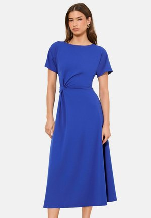 Woman wearing a short-sleeve, knee-length royal blue dress with a side knot detail, standing against a plain white background.