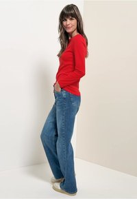 Rotes Langarmhemd, blaue Schlagjeans, hellfarbene Schuhe. Das Modell steht seitlich vor einem neutralen Hintergrund, die Hände in den Taschen.