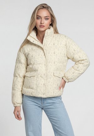 Junge Frau trägt eine hellbeige gesteppte Jacke mit Blumenmuster, hohem Kragen und vorderen Klappentaschen, kombiniert mit hellblauen Jeans, steht mit der Hand auf der Hüfte.