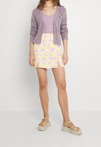 En lavendelfärgad stickad cardigan över en ljuslila topp, tillsammans med en vit kjol med gula och rosa blommönster samt sidofåror. Ljusfärgade platåsandaler.