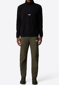 Schwarzer Fleece-Pullover mit einem Viertel-Reißverschluss und weißem Logo auf der Brust, kombiniert mit olivgrünen Cargo-Hosen mit Seitentaschen und Logos.