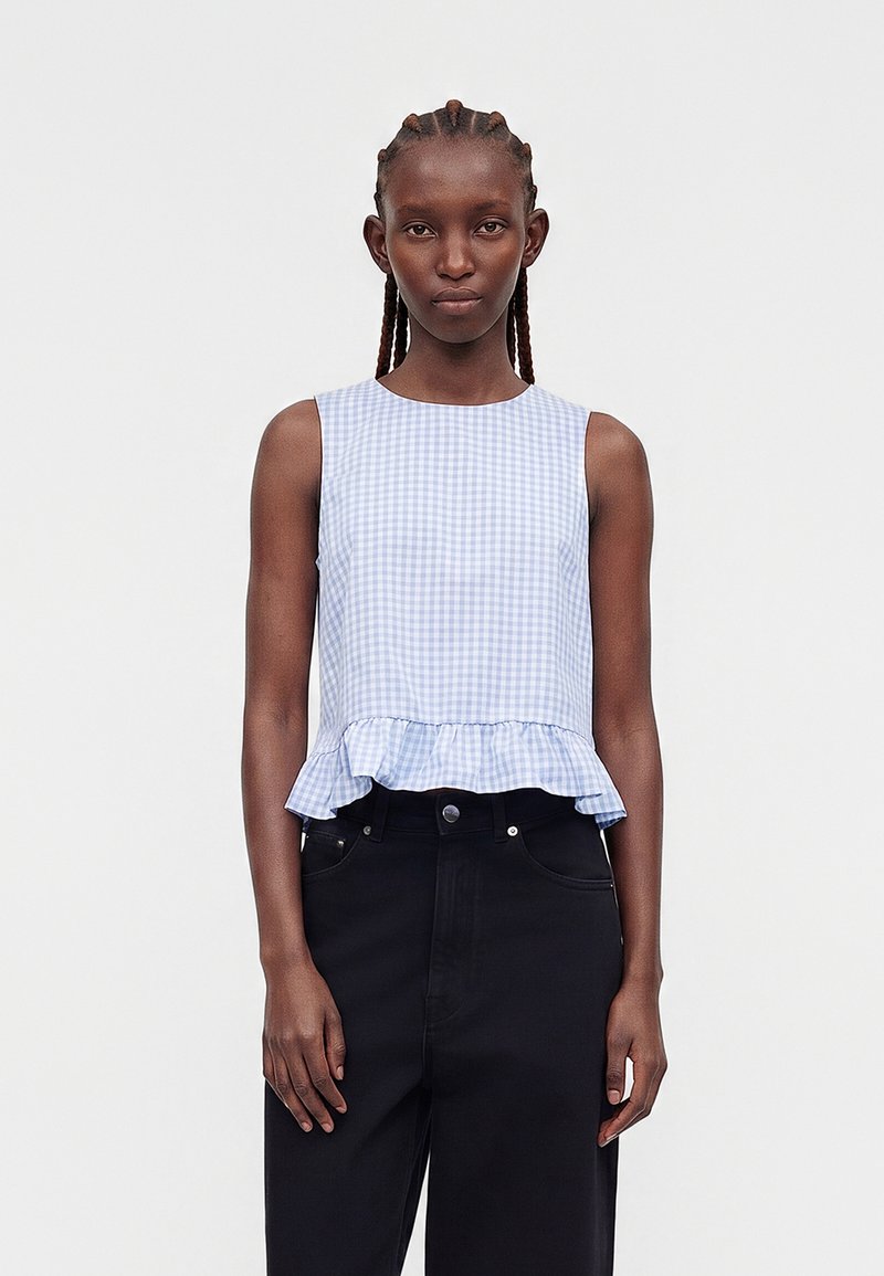 Femme aux cheveux tressés porte un haut sans manches à carreaux bleu et blanc avec un ourlet à volants et un pantalon noir taille haute.
