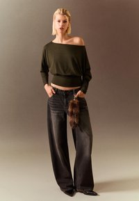 Langærmet, off-the-shoulder, mørkegrøn stribet top med sorte vide jeans, der har en brun pelsdetalje og spidse sorte sko.