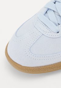 Világoskék velúr sneaker, texturált barna gumitalppal, varrott részletekkel és azonos színű fűzőkkel. Közeli kép az orr részen.