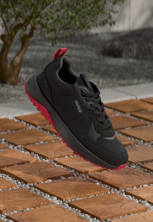 Zwarte sport schoen met een gestructureerde afwerking, voorzien van een rode rubberen zool en zwarte veters, zwevend boven een patroon van stenen.