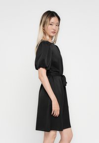 Femme avec de longs cheveux blonds, portant une robe noire longueur genoux avec des manches bouffantes et une ceinture nouée à la taille, se tenant devant un fond blanc uni.