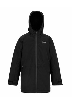 Uśmiechnięty chłopiec w czarnej kurtce parka, oliwkowo zielonym swetrze na zamek, czarnych spodniach typu cargo, torbie na ramię oraz beżowych sportowych butach stoi na tle jednolitego tła.