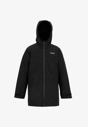 Uśmiechnięty chłopiec w czarnej kurtce parka, oliwkowo zielonym swetrze na zamek, czarnych spodniach typu cargo, torbie na ramię oraz beżowych sportowych butach stoi na tle jednolitego tła.