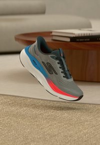Chaussure de course Skechers grise avec des accents bleus et rouges flottant au-dessus d'un tapis beige, d'une table en bois et de livres dans un salon.