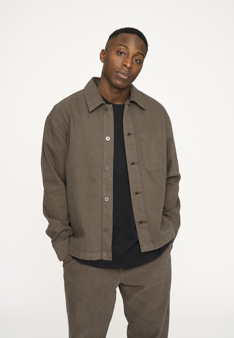 Brun bomulds skjortejakke med en afslappet pasform, knaplukning og brystlomme, parret med mørke bukser. Glat tekstur og minimal dekoration.