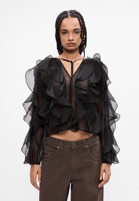 Blusa nera trasparente con volant a strati, maniche lunghe e scollatura a V profonda legata con un cordoncino. Abbinata a jeans marroni.