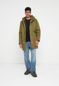 Mann mit olivgrünem Parka mit Fellfutter, schwarzem Shirt, hellblauen Jeans und schwarzen Sneakern, stehend vor weißem Hintergrund.