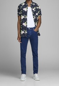 Camicia a maniche corte con stampa floreale scura, t-shirt bianca sotto, jeans blu e sneakers bianche. Tessuto morbido, vestibilità casual.