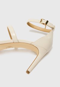 Beige laklæder højhælede sandal med ankelrem, der har en guldfarvet spænde og en glat, blank tekstur.