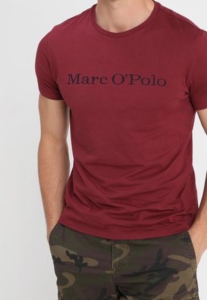 Man wearing a maroon Marc O'Polo t-shirt and green camouflage shorts with hands in pockets against a neutral background.