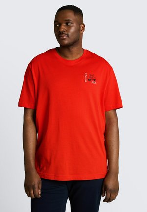 Mann trägt ein hellrotes T-Shirt mit einer kleinen schwarzen Grafik auf der linken Brust und blickt nach rechts vor einem schlichten hellen Hintergrund.