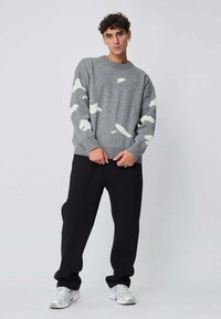 Pull en tricot gris avec des motifs de nuages blancs, col rond et poignets côtelés. Associé avec un pantalon de survêtement noir et des baskets claires.