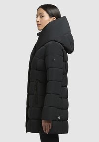 Giacca a vento nera con collo alto, design trapuntato e cappuccio. Presenta una tasca con zip e una piccola etichetta con il marchio vicino all'orlo.