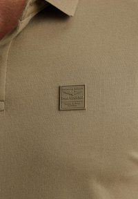 Khaki poloshirt gemaakt van gestructureerde stof met een kraag en een metalen badge met "PME LEGEND" en details over hoogtetesten.