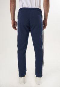 Marina atletiska byxor med vita sidostrick, tillverkade av mjukt tyg. Har elastisk midja och tapered ben-design.