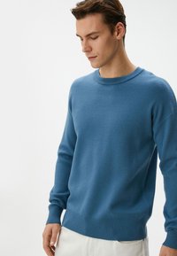 Blauer Strickpullover mit Rundhalsausschnitt, gerippten Bündchen und Saum. Weiche Textur mit taillierter Passform. Getragen mit hellen Hosen.