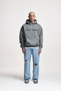 Siv hoodie sa kapuljačom s prednjim džepom i logotipom "ZY STUDIOS", u kombinaciji sa svijetloplavim iznositim trapericama i crno-bijelim tenisicama.