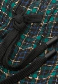 Primer plano de un cordón negro trenzado atado en un nudo sobre una tela de cuadros verde, azul, amarillo y negro.