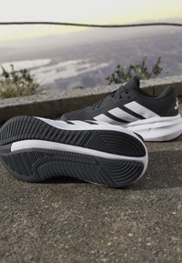 Chaussures de running noir et blanc avec semelle texturée posées sur un trottoir extérieur près d'une barrière en pierre avec un paysage urbain flou en arrière-plan.