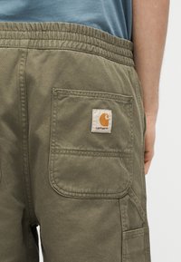 Κοντινό πλάνο σε χακί παντελόνι με ελαστική μέση και μπαλώμα με το λογότυπο Carhartt στην πίσω τσέπη, το οποίο φοριέται με μπλε πουκάμισο.