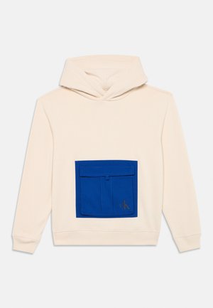 Kremowy bawełniany hoodie z dużą niebieską kieszenią z przodu. Kieszeń ma klapkę i wytłoczone logo w złotym kolorze. Miękka tekstura i luźny krój.