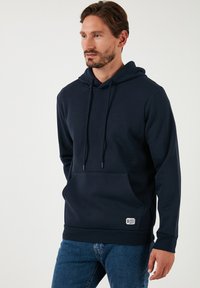 Felpa con cappuccio blu navy realizzata in tessuto morbido, dotata di tasca a marsupio, cordini regolabili e una piccola patch con logo sull'orlo.